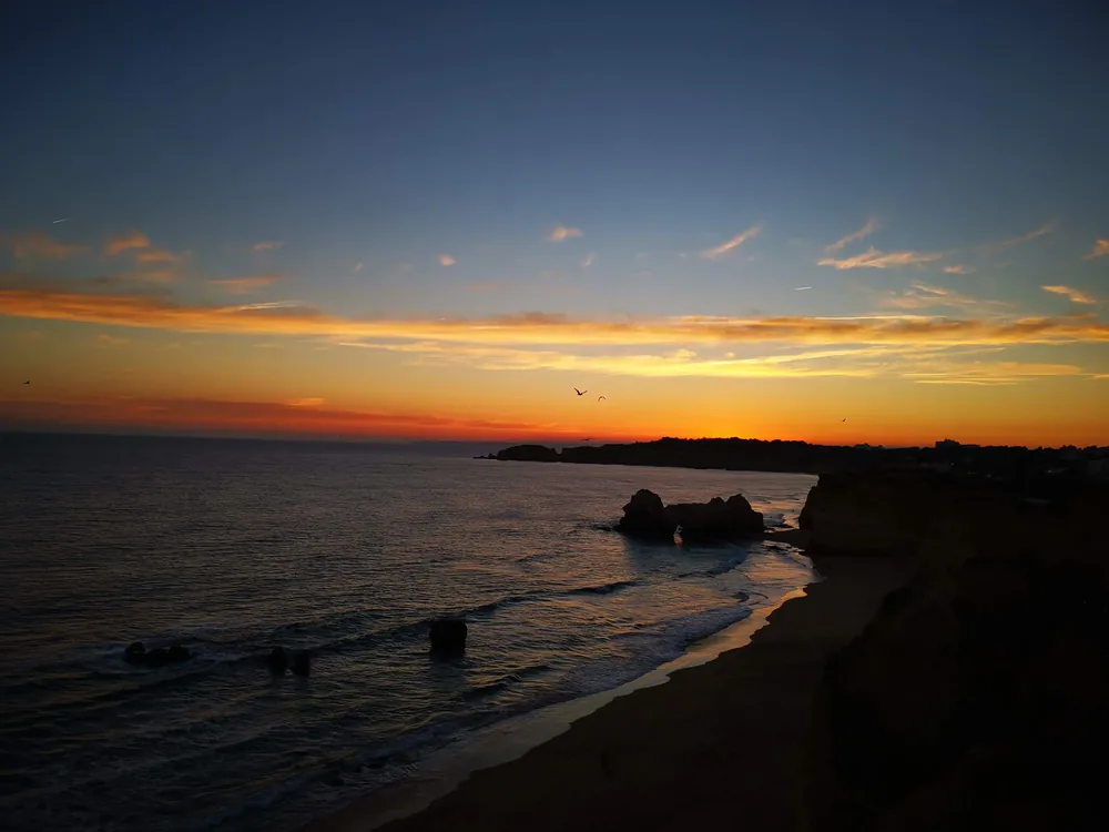 Sunset over the Atlantic from a clifftop, orange and gold sky streaked with cloud, dark rock stacks and a headland in silhouette, and small waves washing onto the sand below.