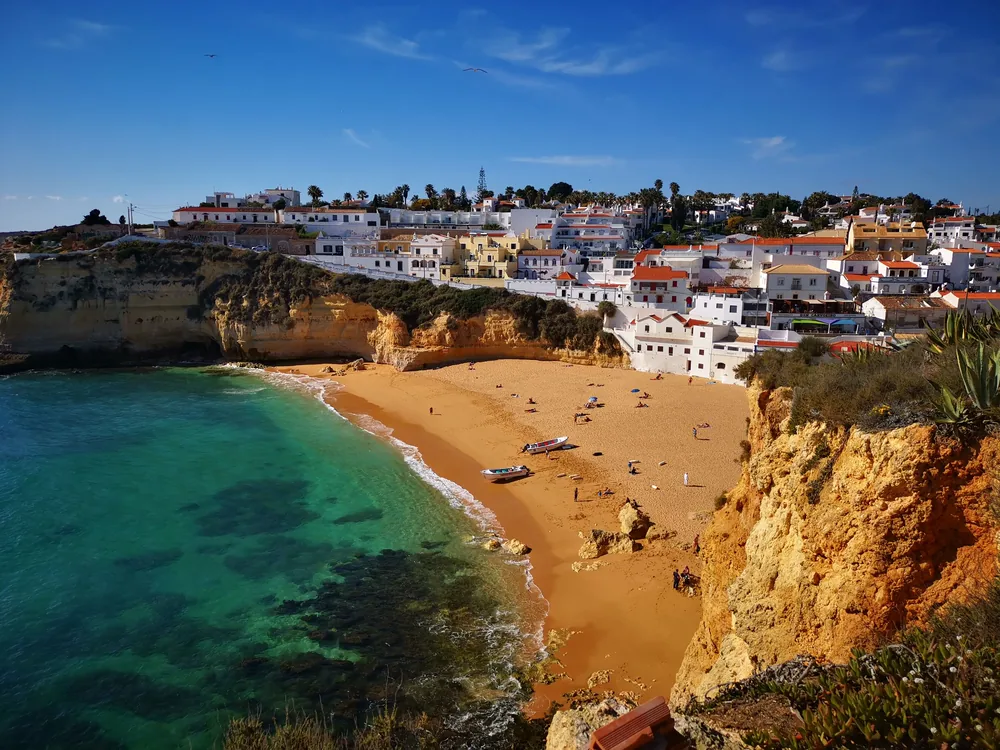 A high view over a golden crescent beach with clear turquoise water, orange eroded cliffs, small boats on the sand and a whitewashed coastal town with palm trees on the headland above.