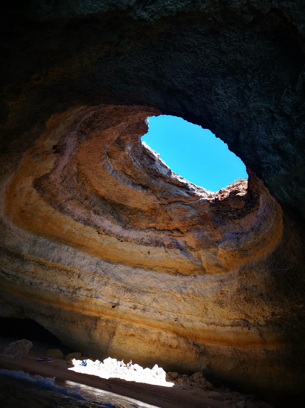 Inside the Benagil sea cave, looking up through the circular opening in the ceiling to blue sky, with layered golden and grey limestone walls curving around and a sliver of sunlit beach far below.