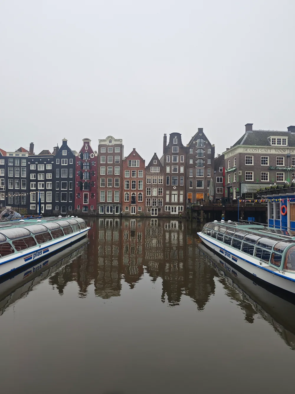 Two glass-roofed canal tour boats moored on a calm canal, their reflections and a row of tall narrow gabled canal houses mirrored in the still water.