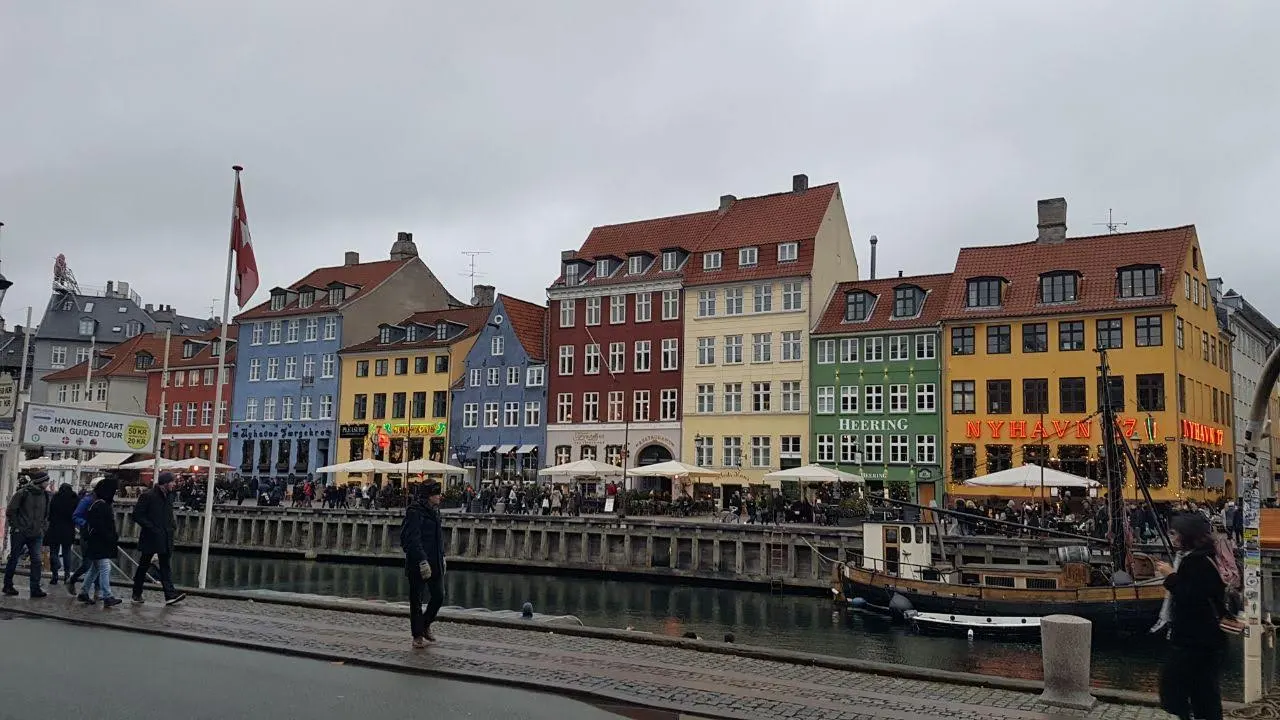 The colourful gabled townhouses of Nyhavn in blue, yellow, red and green with red tile roofs, a Danish flag flying, a wooden boat moored at the quay and people walking along the canal.