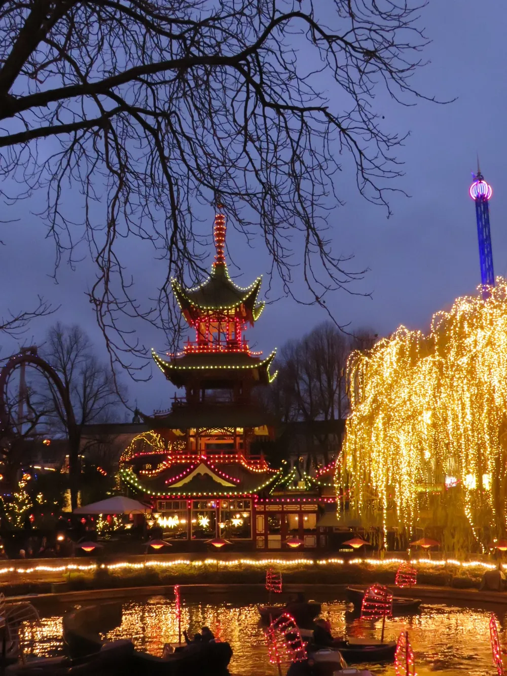 Tivoli Gardens after dark, with a Chinese-style pagoda tower outlined in red and green lights, bare branches across the top of the frame and a tree draped in golden lights, boats on the canal below.