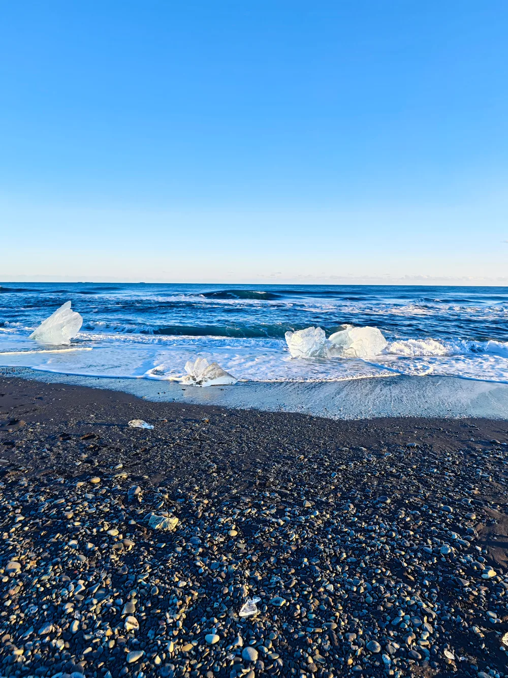 Chunks of clear blue ice washed up on a black sand beach as waves roll in from the Atlantic under a deep blue sky.
