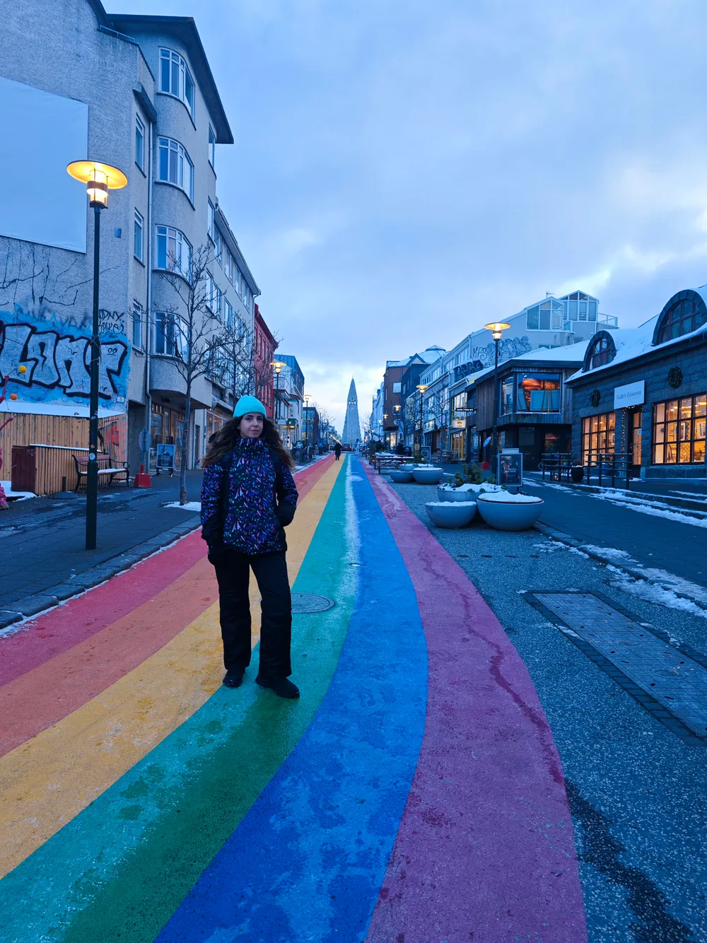 A traveller in a teal beanie standing on the rainbow-painted crossing of Skólavörðustígur in Reykjavík, with Hallgrímskirkja's spire rising at the end of the street.