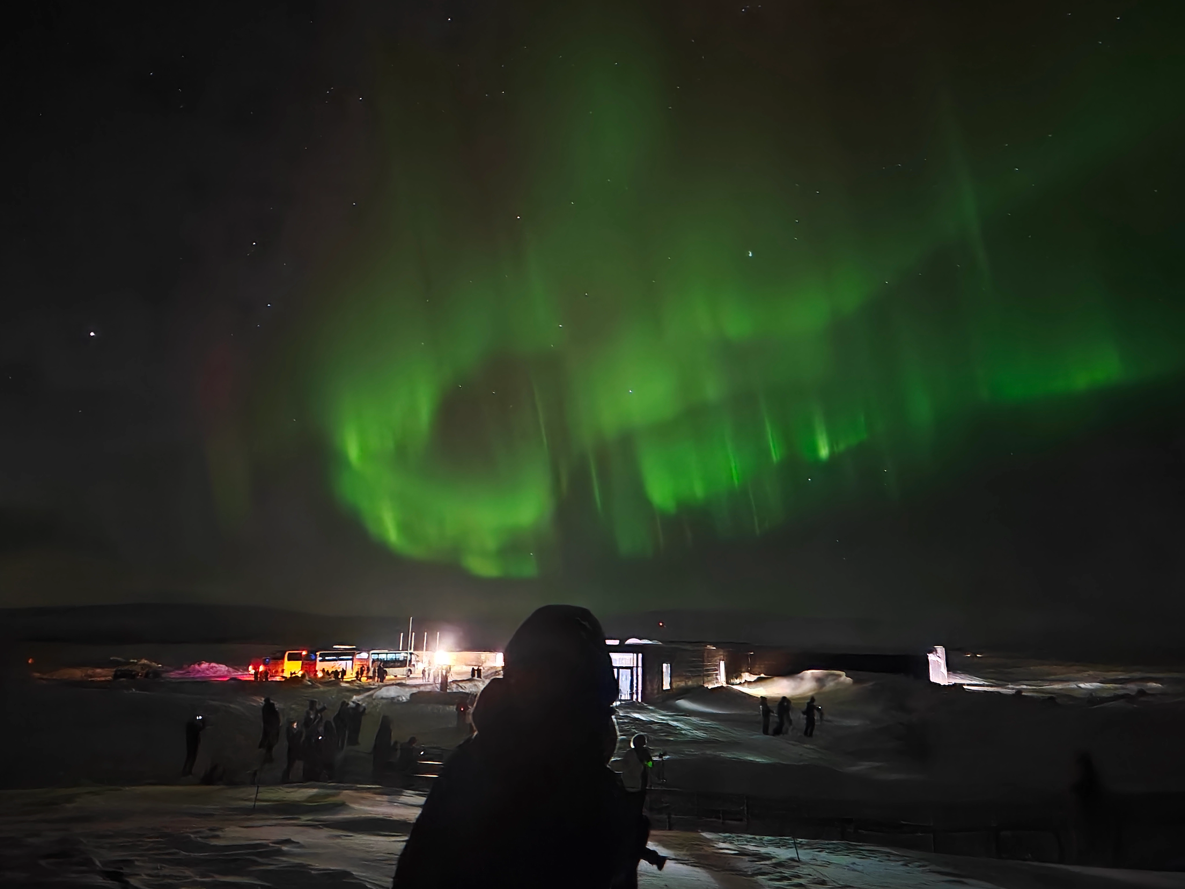 Curtains of bright green aurora swirling across a starry sky above a snow-covered town, with a hooded figure watching from a frozen field.