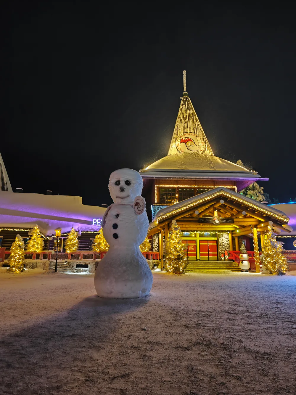 A three-tier snowman lit up at night in front of the golden-lit peaked roof and wooden entrance of Santa Claus Village.