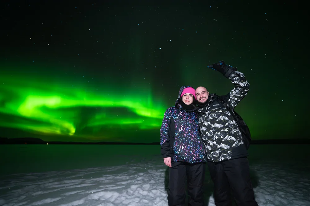 Two travellers in insulated suits standing on a snowy clearing under a starry sky, one arm raised in front of vivid green aurora ribbons.