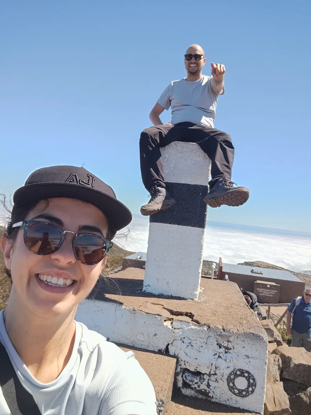 Two travellers at a summit marker on Pico do Arieiro with a sea of clouds below