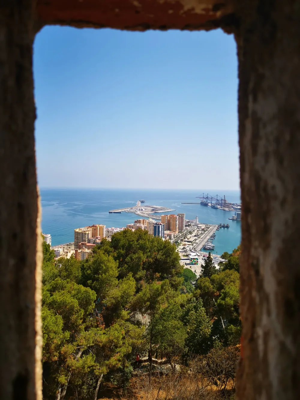 The port of Málaga seen through a weathered stone window opening, with the marina, a docked cruise ship and apartment blocks below and the blue Mediterranean beyond.