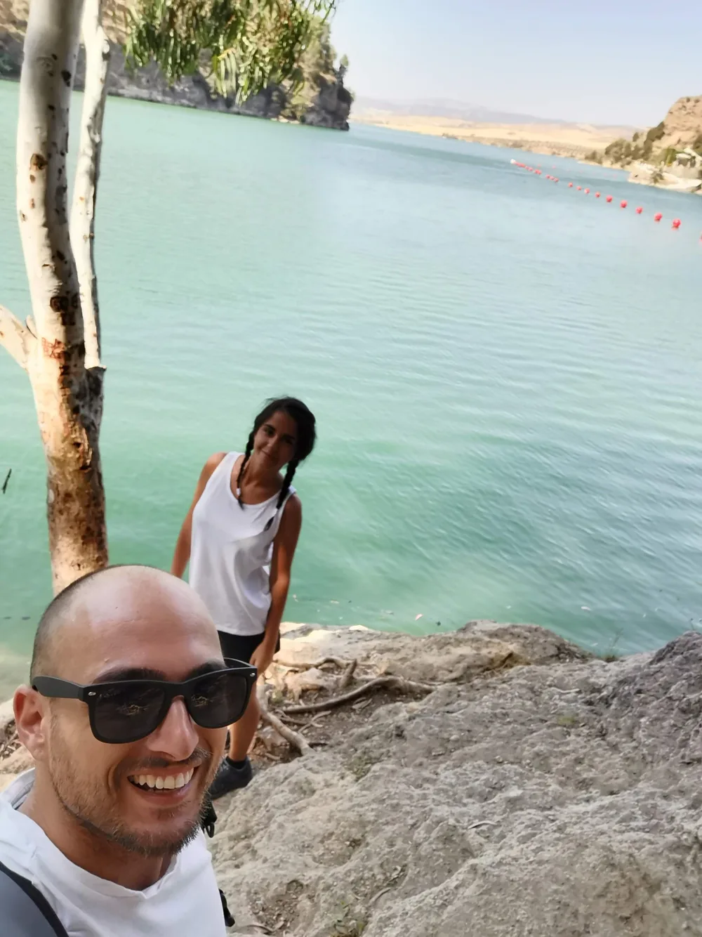Two travellers smiling on a rocky bank above milky turquoise water, with a line of red buoys and a reed-fringed shore across the reservoir.