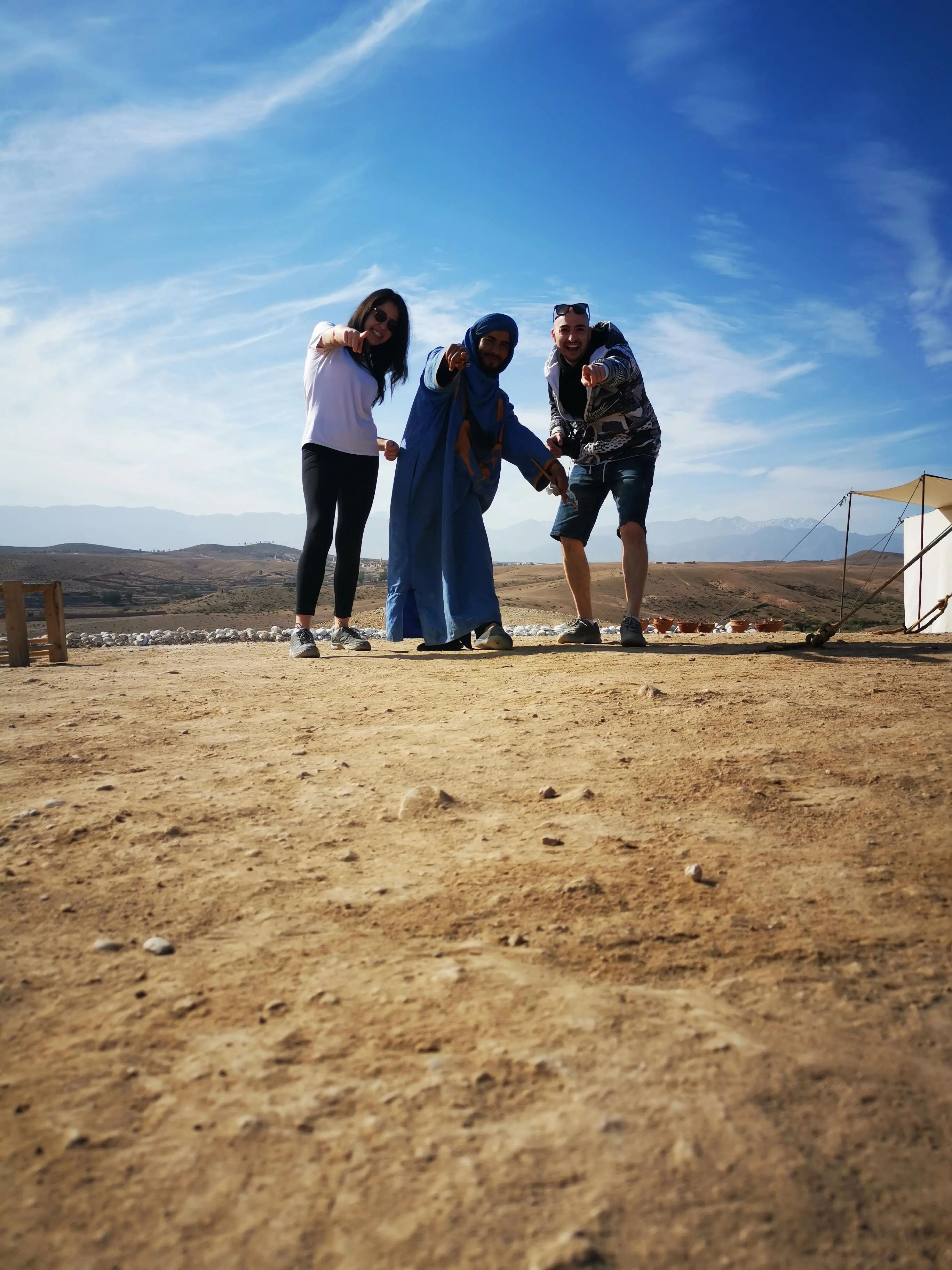 Two travellers pointing at the camera beside a blue-robed local, standing on a dusty launch field with the snow-dusted Atlas Mountains behind and a deep blue sky above.