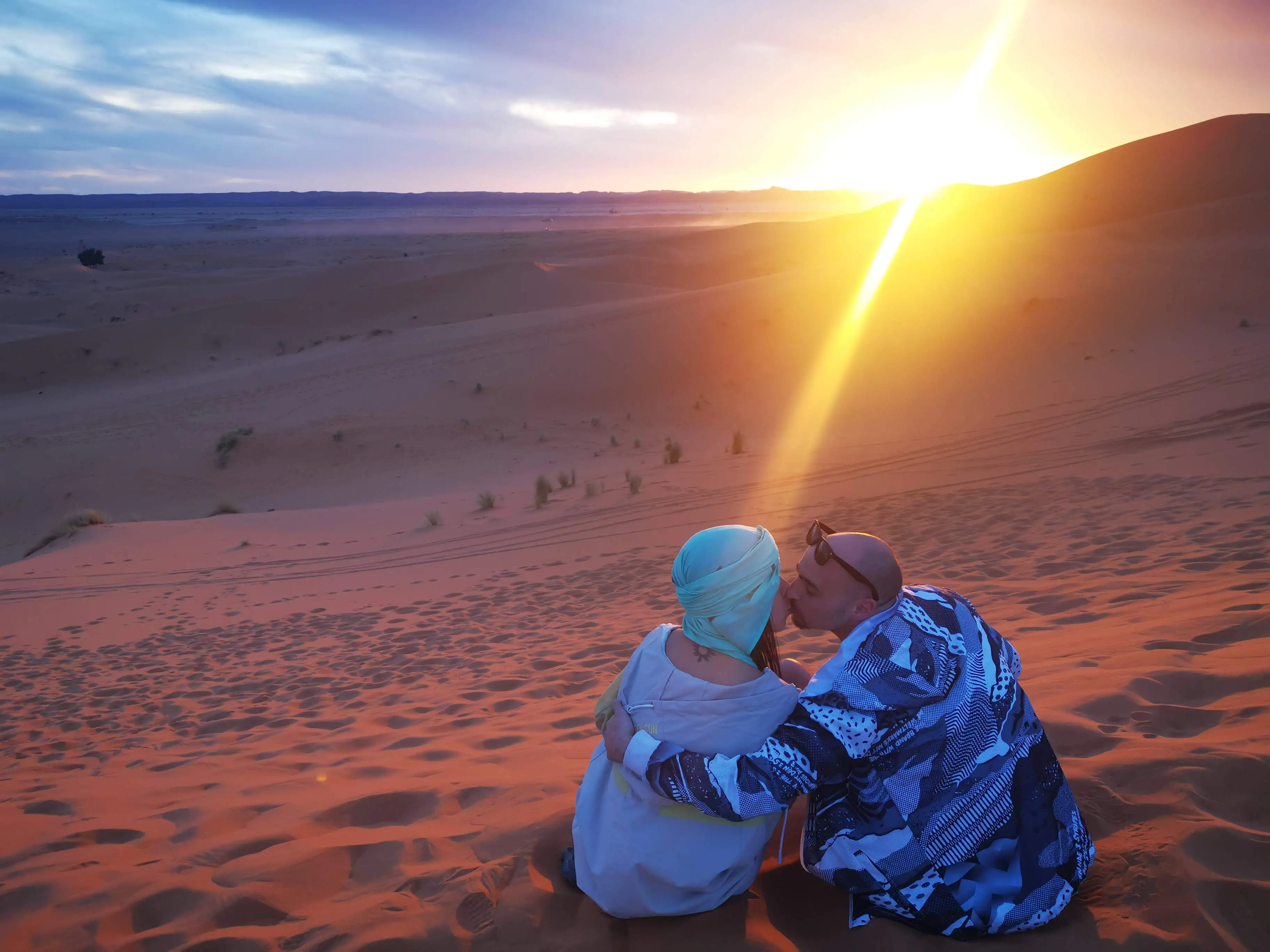 A couple sitting close together on red desert dunes as the low sun throws long shadows across the sand and a dark dune ridge behind.