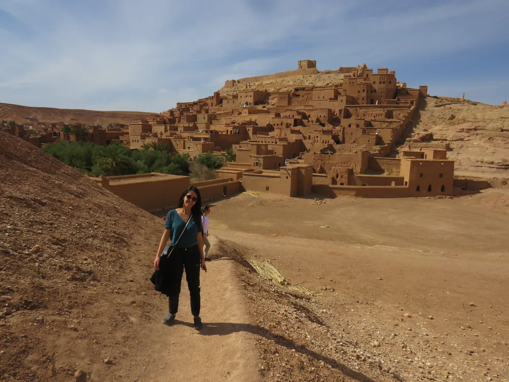 A vast ochre earthen kasbah rising in terraces up a barren hillside, with a palm grove and dry brown mountains stretching away behind.
