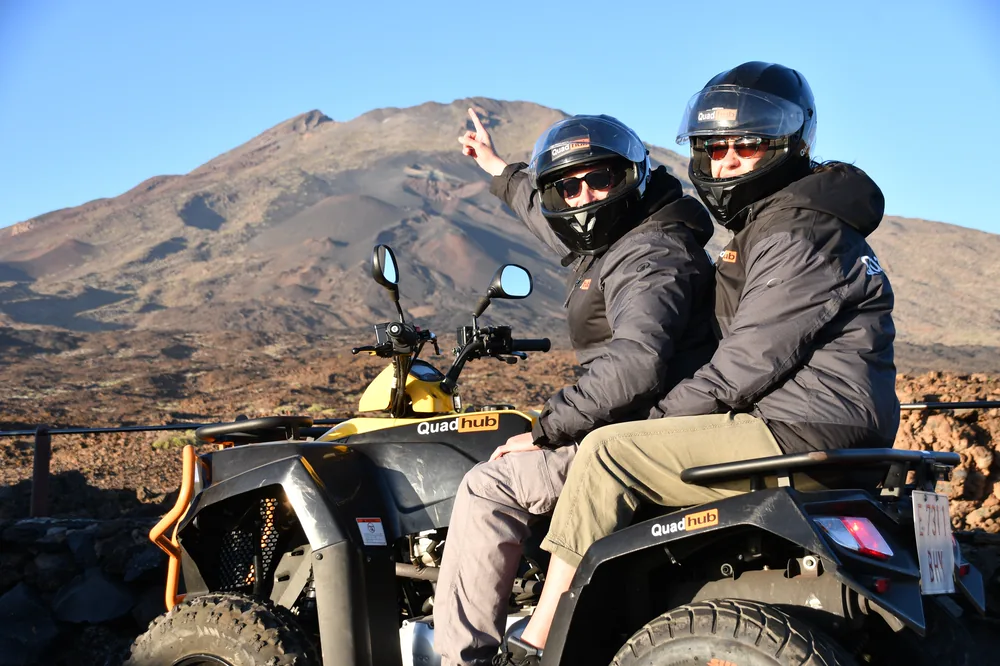 Two helmeted travellers on a quad bike parked on black volcanic rock, pointing towards a bare brown mountain under a clear blue sky.