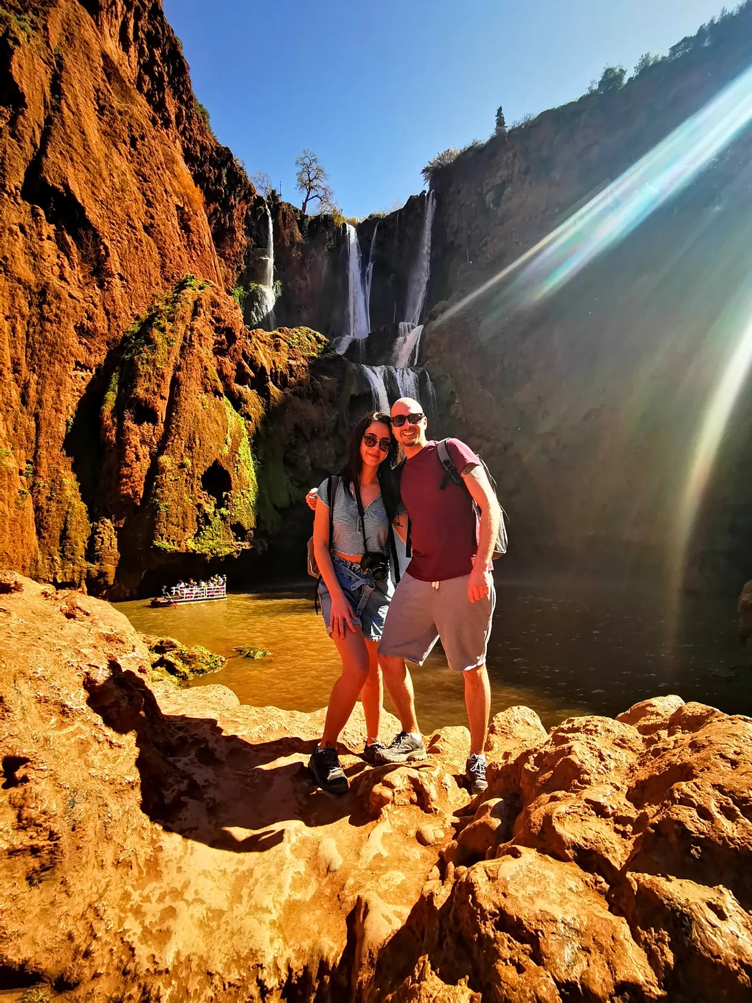 Valentina and Ivan on a sunlit rocky path beside a waterfall